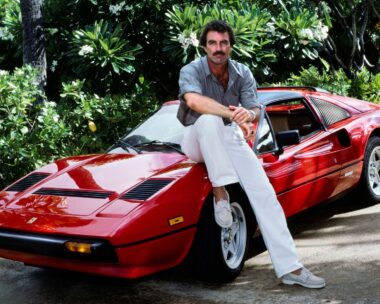 Tom Selleck sits on red car for a Magnum PI shoot. He's wearing white pants and a Hawaiian button-up shirt.