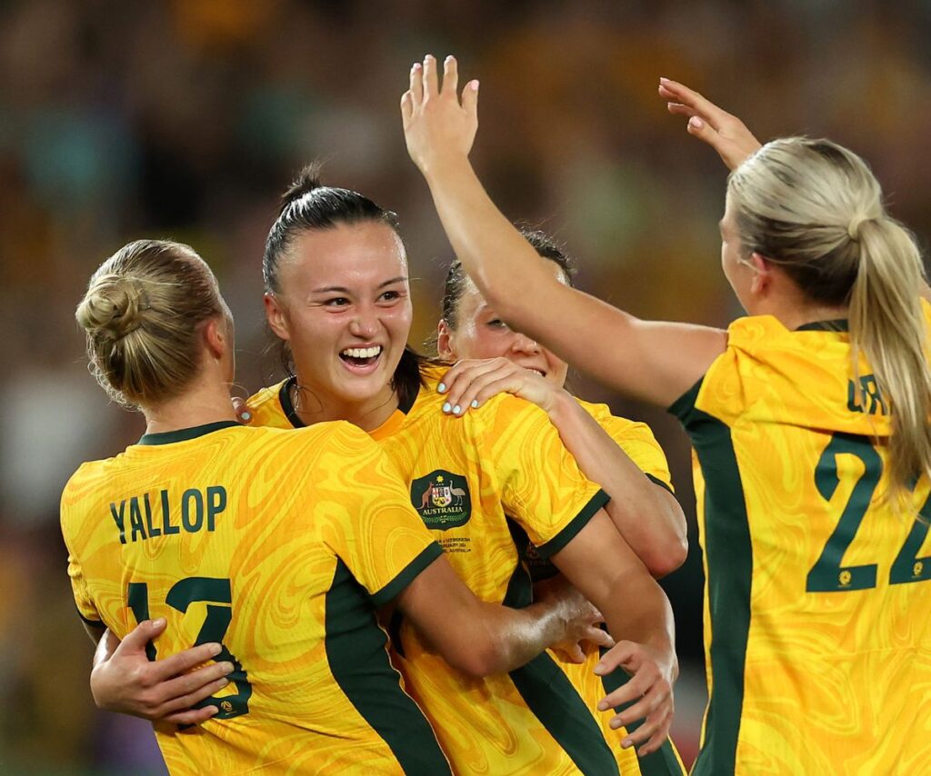 Amy Sayer smiles as the Matildas team hugs, celebrating a likely goal