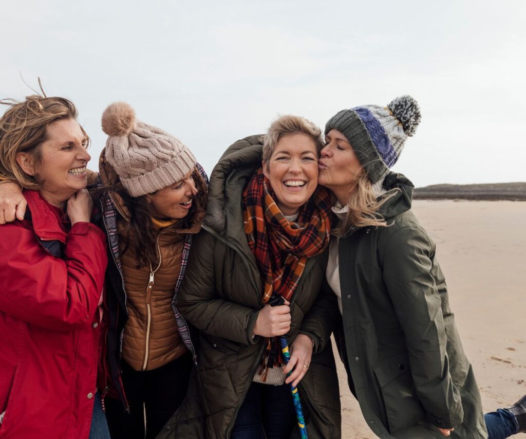 Mid-aged female friends on the beach, dressed in winter coats, scarfs and hats look happy to be together
