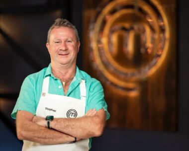 MasterChef Stephen Dennis crosses arms in front of MasterChef logo wearing white apron and blue collared shirt