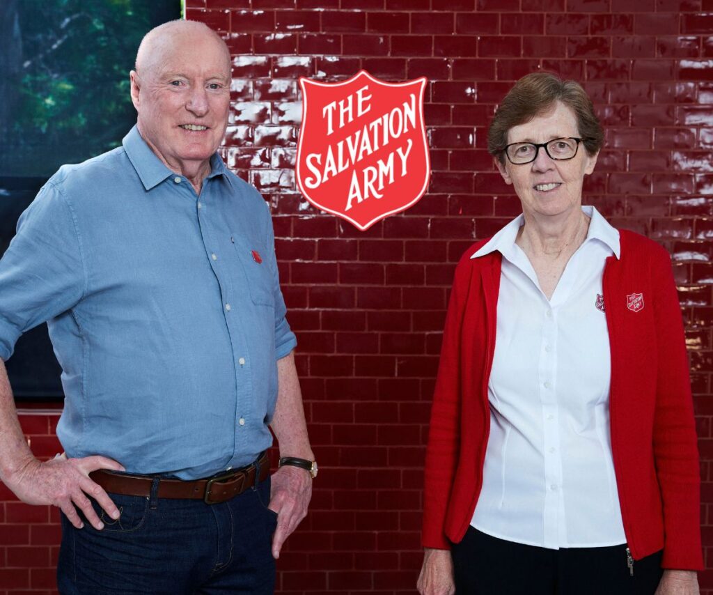 Ray Meagher and Salvos' Chief Miriam Gluyas smile in front of red brick wall with salvation army emblem between them