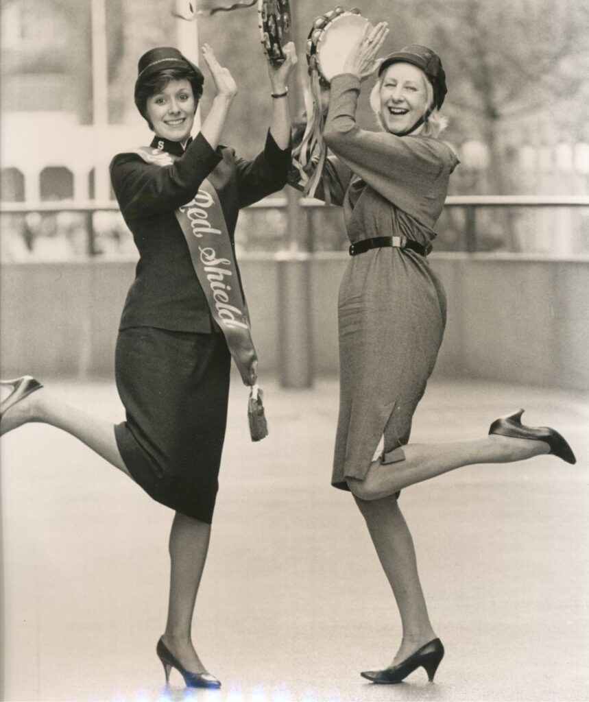 A nostalgic black-and-white image of the first Red Shield, with two women smiling and kicking up their heels, hitting tambourines