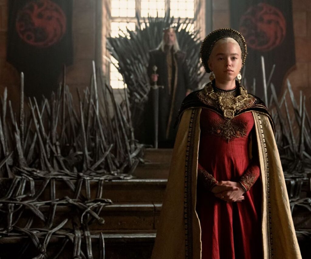 In Westeros, Rhaenyra wears a read gown with gold cape, standing in front of the Iron Throne in filming for House of the Dragon