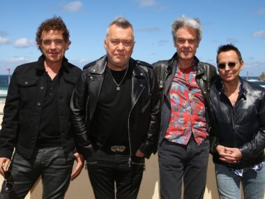 Cold Chisel band stands outside in black jackets against a beach backdrop