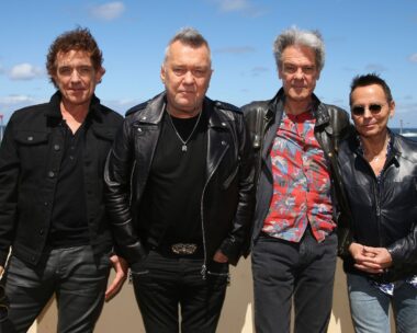 Cold Chisel band stands outside in black jackets against a beach backdrop