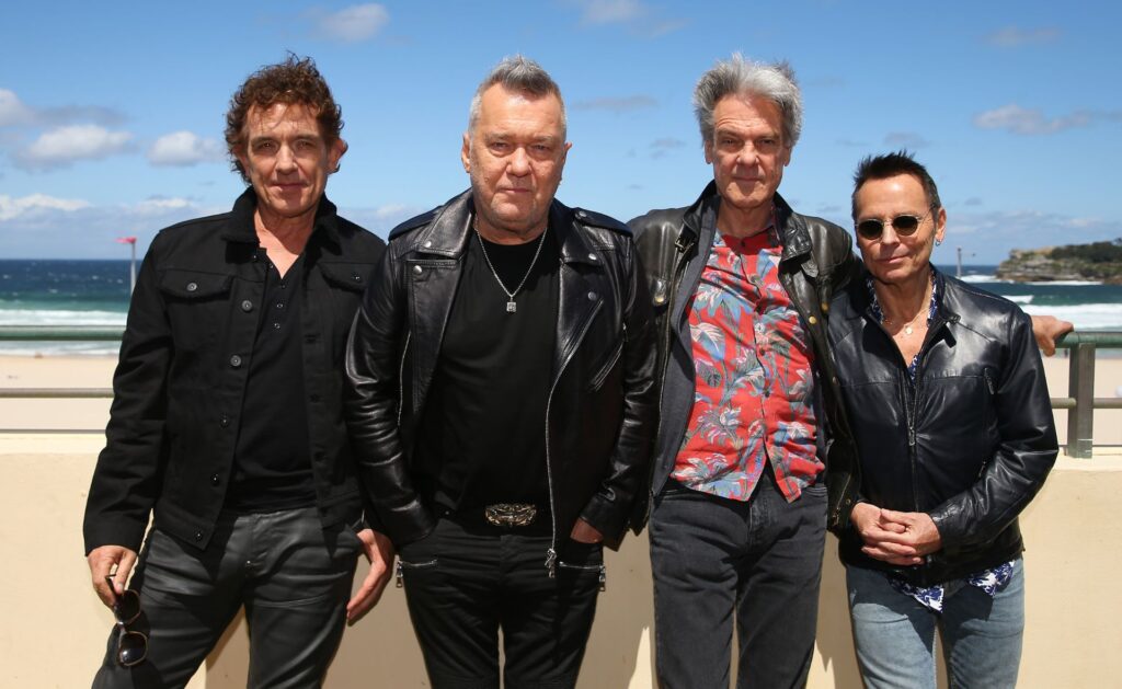 Cold Chisel band members stand in front of the beach looking at the camera in black jackets