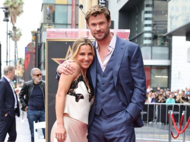 Elsa wearing a white dress with black lace trim stands with husband Chris Hemsworth, wearing blue suit with vest, smiling