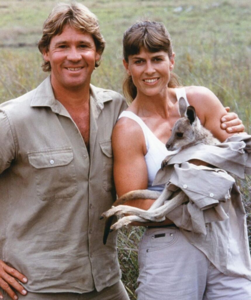 Terri and Steve Irwin smile at camera, Terri holding a baby kangaroo and Steve with his arm around Terri