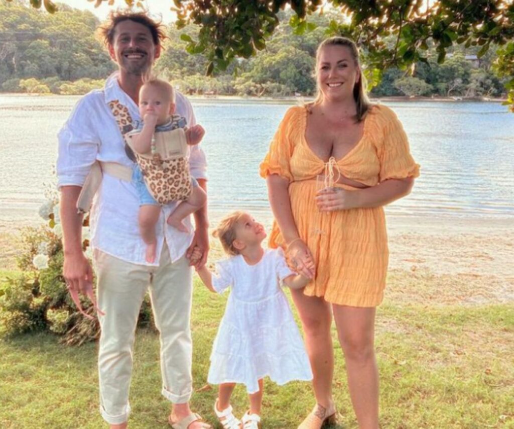 Dream Home winner Rhys Almond stands with his family, holding his newborn in a baby carrier on his chest, while holding his little girl's hand