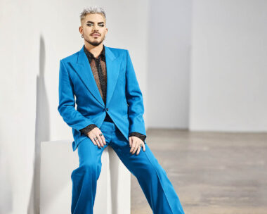 Adam sits on a stool in a two-piece blue suit.