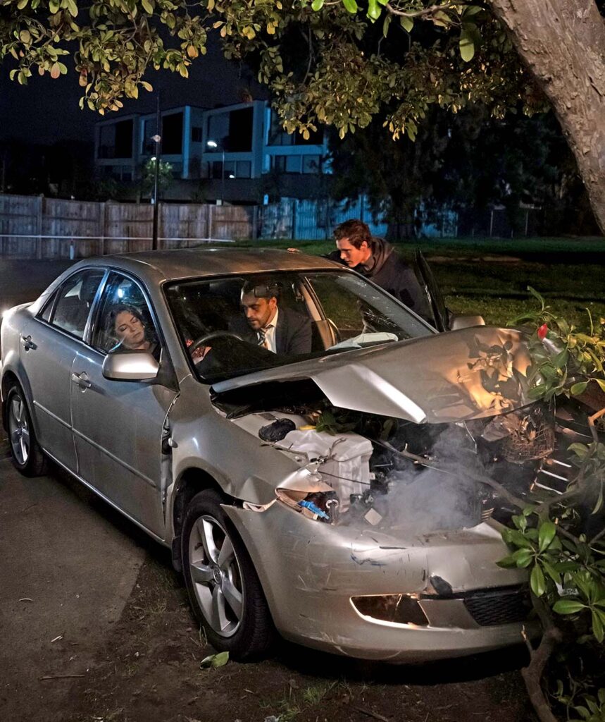 The car has crashed into a tree and is severely damaged. Terese and Andrew are in the front seats. Terese is injured and Andrew checks on her.